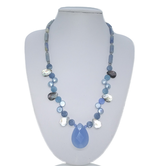 A beaded statement necklace in shades of blue, gray, and white, featuring a blue teardrop pendant, displayed on a white jewelry bust.