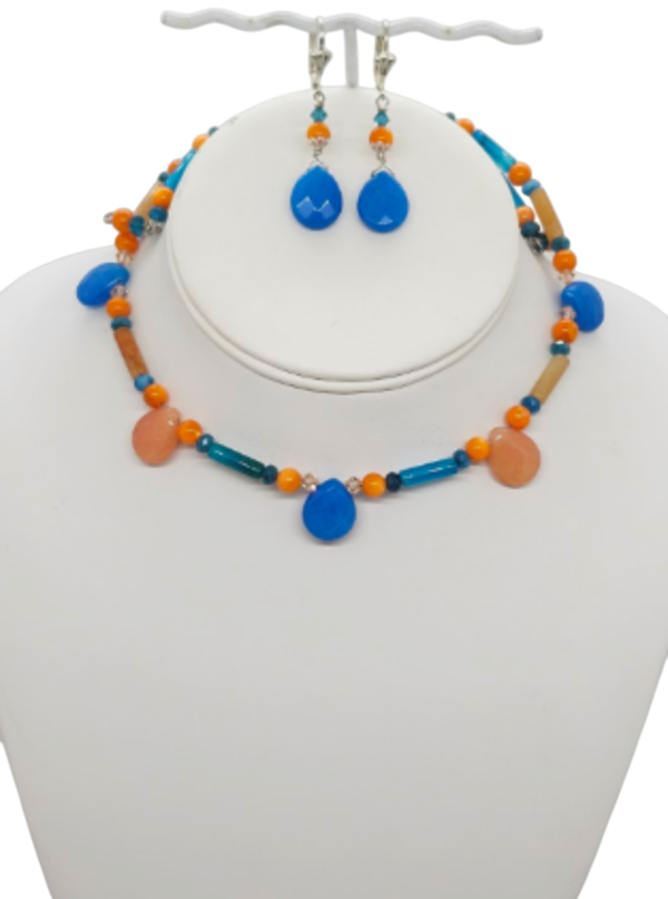 A jewelry set displayed on a white mannequin bust, featuring a beaded choker-style necklace and matching drop earrings. The set includes bright turquoise blue teardrop beads, orange round beads, teal blue cylindrical beads, and orange crystals.