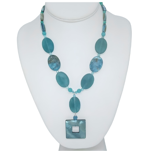 Teal blue, seafoam green and turquoise gemstone statement necklace on a white display bust. The necklace features smooth oval jade-style beads, patterned blue crazy lace agate beads, and a square, iridescent teal-blue Mother of Pearl pendant.