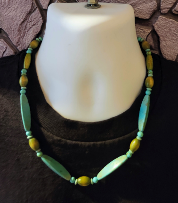 A beaded necklace displayed on a white mannequin bust wearing a black top, featuring large turquoise bar beads and olive green oval beads in addition to smaller round and rondelle turquoise beads.