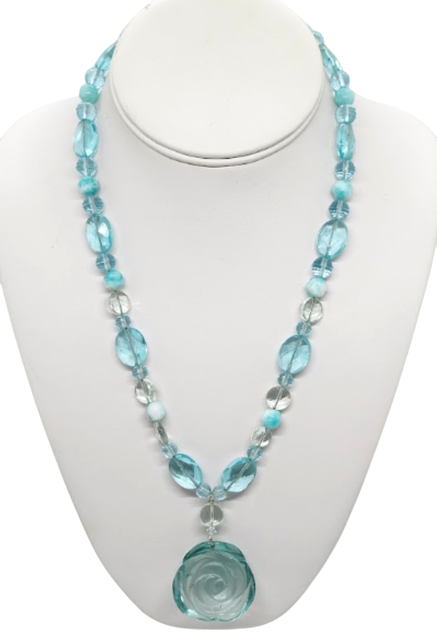 A beaded necklace with light aqua green and clear faceted beads, featuring a large carved light aqua green rose pendant, displayed on a white mannequin bust.