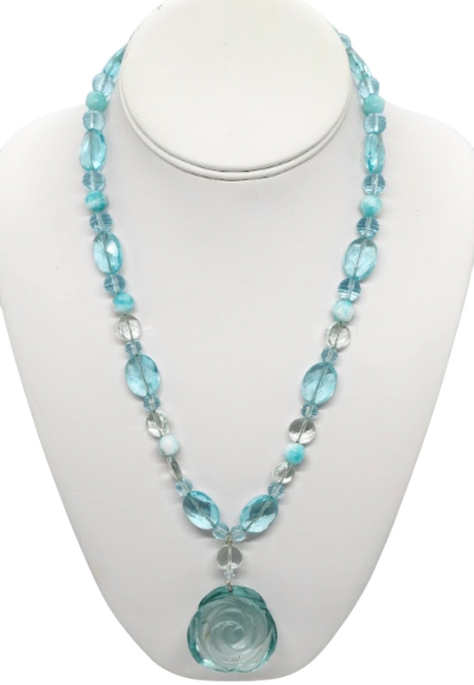 A beaded necklace with light aqua green and clear faceted beads, featuring a large carved light aqua green rose pendant, displayed on a white mannequin bust.