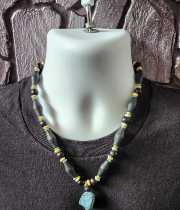 A beaded necklace displayed on a white mannequin bust wearing a black t-shirt, featuring black, beige and aqua wood beads and a turquoise blue nugget stone pendant.