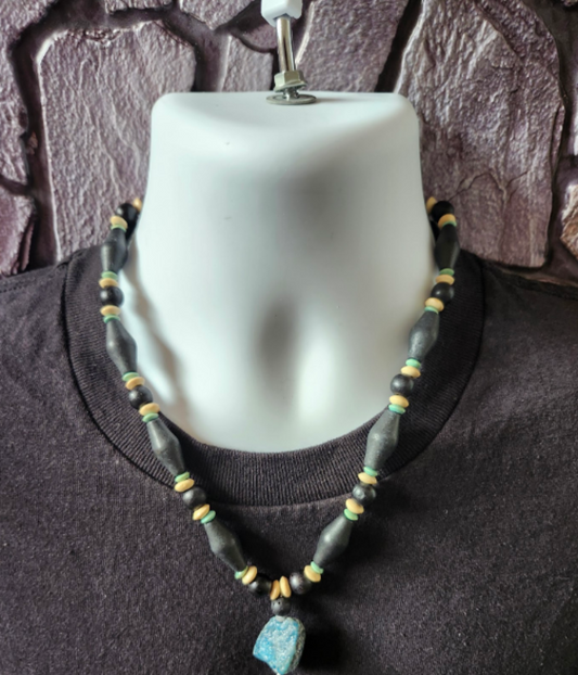 A beaded necklace displayed on a white mannequin bust wearing a black t-shirt, featuring black, beige and aqua wood beads and a turquoise blue nugget stone pendant.