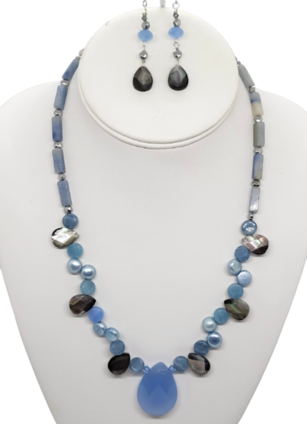 A jewelry set displayed on a white mannequin bust, featuring a statement necklace and matching  blue and gray teardrop drop earrings. The jewelry is made of various blue semi-precious stones and gray semi-precious stones and shells.  The necklace has a medium-sized faceted blue chalcedony  teardrop pendant.