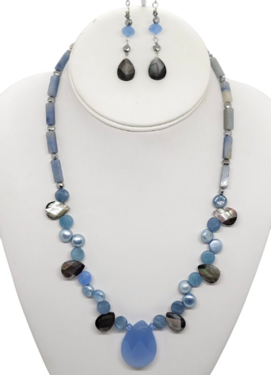 A jewelry set displayed on a white mannequin bust, featuring a statement necklace and matching  blue and gray teardrop drop earrings. The jewelry is made of various blue semi-precious stones and gray semi-precious stones and shells.  The necklace has a medium-sized faceted blue chalcedony  teardrop pendant.