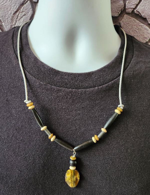 A beaded leather cord necklace displayed on a white mannequin torso wearing a dark gray T-shirt. The necklace features a light gray leather cord, black horn pipe tube beads, small beige and brown wood disc beads, and a  tiger eye stone pendant.