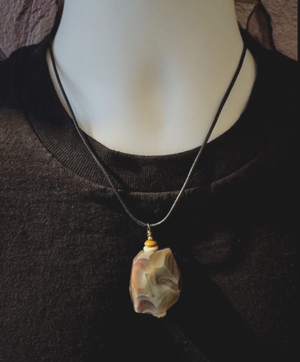 A close-up of a necklace displayed on a white mannequin torso wearing a black top. The necklace has a black leather cord and a polished irregular Botswana agate pendant in beige, cream, gray and brown.