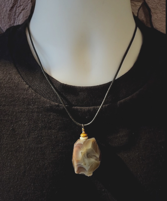 A close-up of a necklace displayed on a white mannequin torso wearing a black top. The necklace has a black leather cord and a polished irregular Botswana agate pendant in beige, cream, gray and brown.