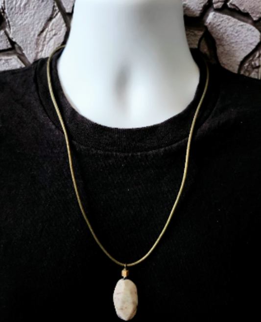 A gold chain necklace with a natural, irregularly shaped light gray stone pendant, displayed on a white mannequin torso wearing a black shirt against a textured stone wall background.