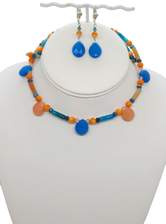 A jewelry set displayed on a white mannequin bust, featuring a beaded choker-style necklace and matching drop earrings. The set includes bright turquoise blue teardrop beads, orange round beads, teal blue cylindrical beads, and orange crystals.  