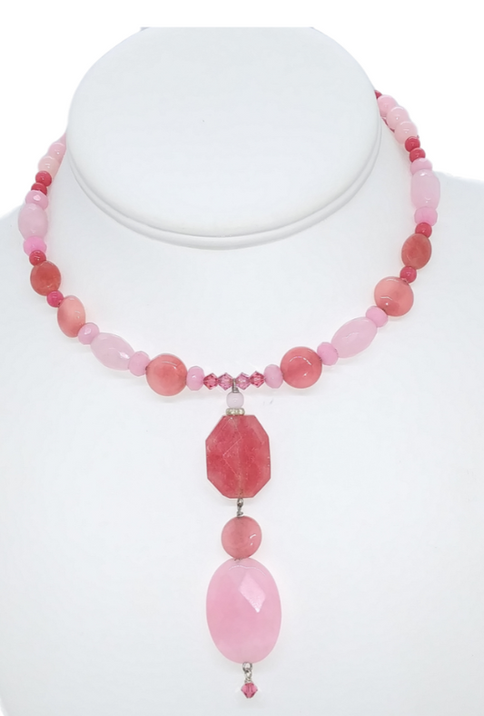 A pink and raspberry-colored beaded necklace displayed on a white mannequin bust. The necklace features a mix of light pink, medium pink, and darker rose pink beads, with a central cluster of small faceted pink beads and a large octagonal pink stone pendant hanging below, followed by a small pink bead and a larger oval pink stone.
