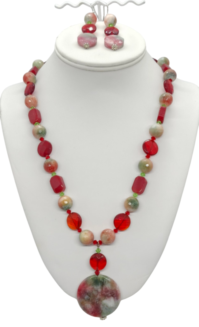 A red, green, peach and beige beaded necklace and matching drop earrings displayed on a white jewelry bust. The necklace features a mix of semi-precious stones and crystals, with a large round multi-colored jade pendant at the center.