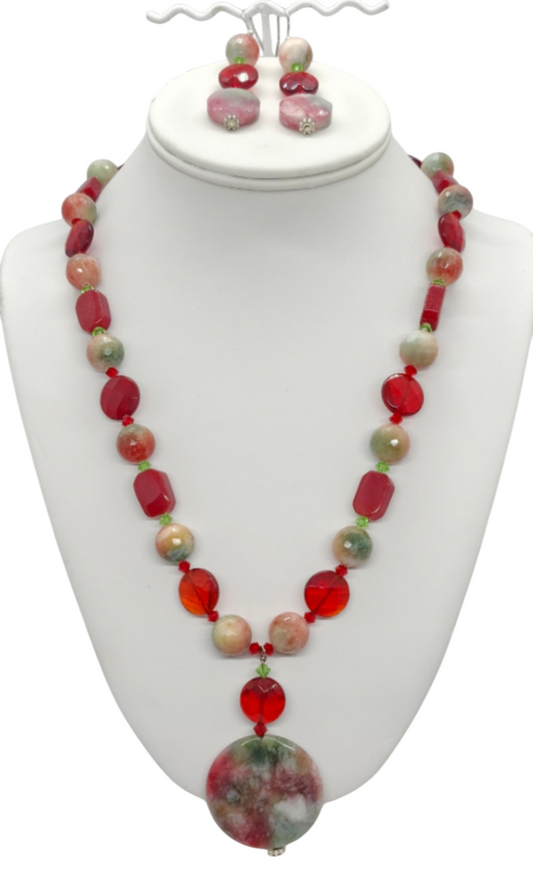 A red, green, peach and beige beaded necklace and matching drop earrings displayed on a white jewelry bust. The necklace features a mix of semi-precious stones and crystals, with a large round multi-colored jade pendant at the center.