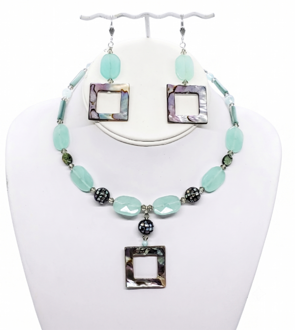 A handcrafted jewelry set featuring a choker necklace and matching drop earrings on a white display stand. The set features smooth, light seafoam green gemstone beads, dark iridescent mosaic shell spheres, and square, open-center mother-of-pearl pendants that reflect aqua, teal, gold, and green hues.